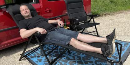 Person in schwarzem T-Shirt und Jeansshorts liegt in klappbarem Liegestuhl neben einem roten Van auf blau gemustertem Teppich; leerer Stuhl und Flasche sichtbar