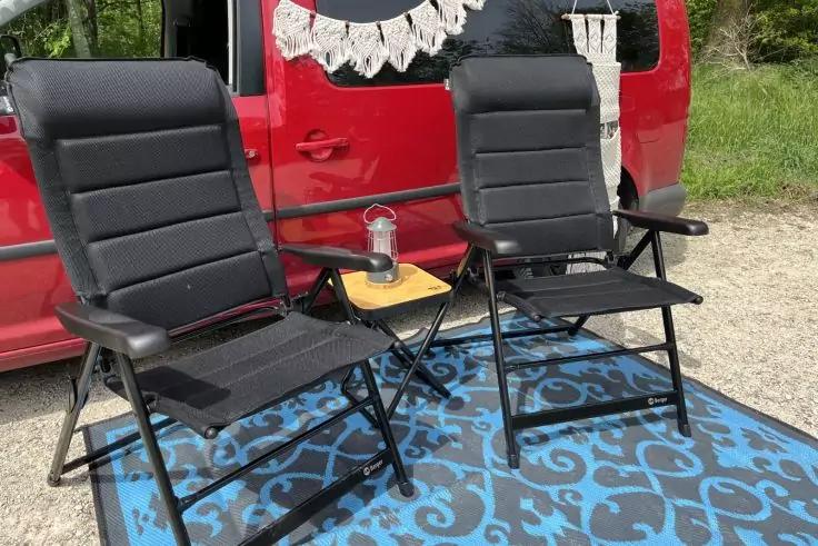 Zwei schwarze Klappst&uuml;hle mit Armlehnen auf blau gemustertem Teppich, dazwischen kleiner Holz-Beistelltisch mit Trinkflasche; im Hintergrund roter Van, wei&szlig;er Makramee-Wandbehang und Kiesboden