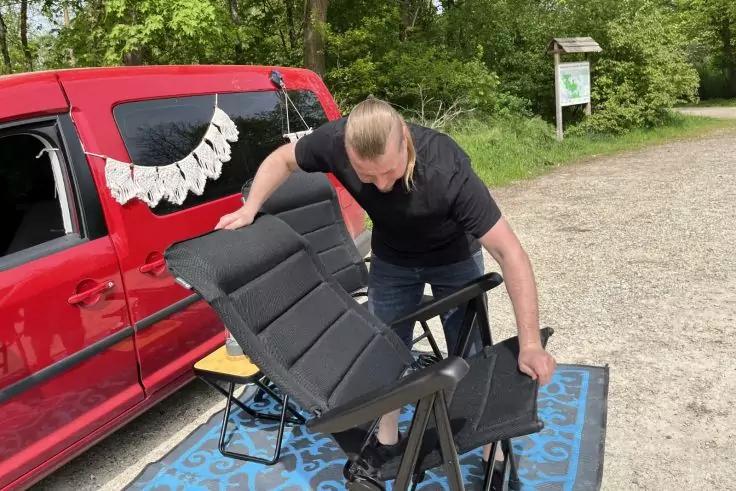 Person beugt sich &uuml;ber einen schwarzen Klappstuhl auf einer blauen Matte neben einem roten Van mit wei&szlig;er Wimpelkette; Kiesweg, B&auml;ume und eine Informationstafel im Hintergrund