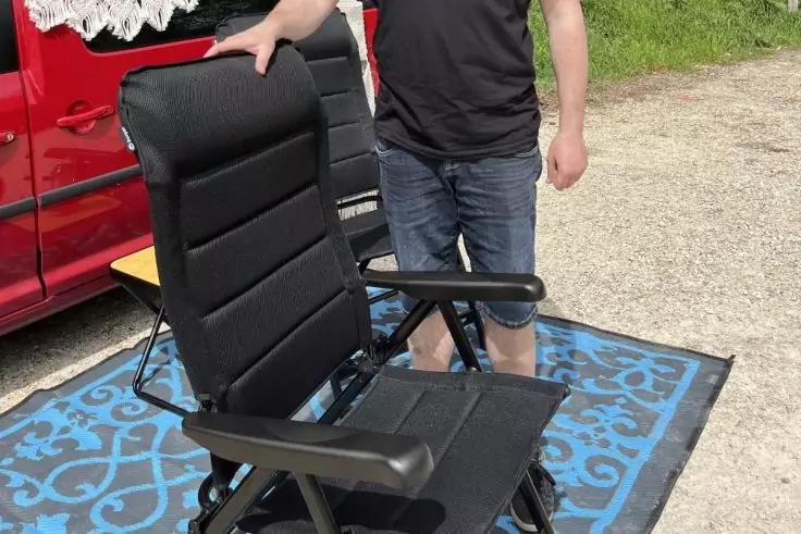Schwarzer Klappstuhl auf blau gemusterter Matte, stehende Person neben dem Stuhl und rotes Fahrzeug im Hintergrund