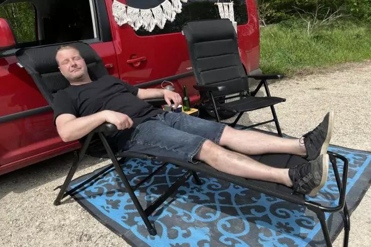 Mann in einem Liegestuhl mit ausgestreckten Beinen auf einer Fu&szlig;st&uuml;tze, neben einem roten Van auf einer blauen, gemusterten Matte; ein weiterer Klappstuhl, Schotterboden und Gr&uuml;n im Hintergrund