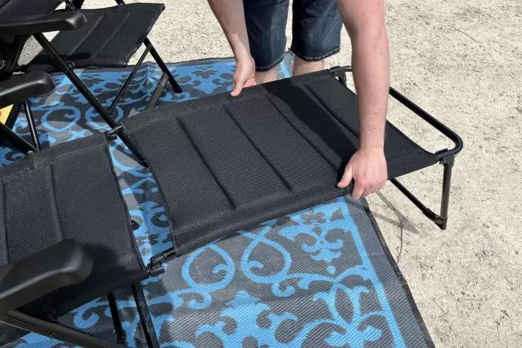 H&auml;nde und Beine einer Person in blauen Jeansshorts, die eine schwarze Sitzfl&auml;che im Rahmen eines Klappstuhls h&auml;lt; der Stuhl steht auf einer blau gemusterten Matte, sandiger Untergrund im Hintergrund