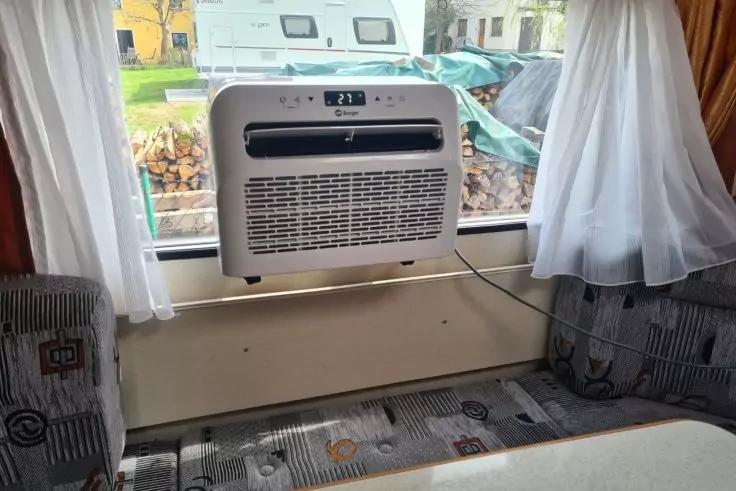 Fenster mit wei&szlig;en Vorh&auml;ngen und in der &Ouml;ffnung eingesetztem Klimager&auml;t, gepolsterte Sitzbank mit gemustertem Stoff darunter, Tischkante im Vordergrund, Holzstapel und geparkte Fahrzeuge drau&szlig;en