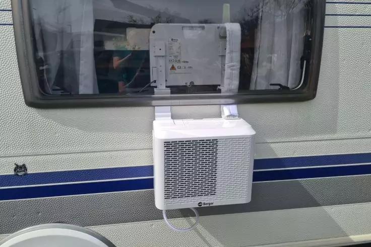 Wei&szlig;es Ger&auml;t mit L&uuml;ftungsgitter unter einem Fenster an der Au&szlig;enwand eines Wohnwagens, Kabel f&uuml;hrt ins Fahrzeug, Gardinen im Fenster sichtbar