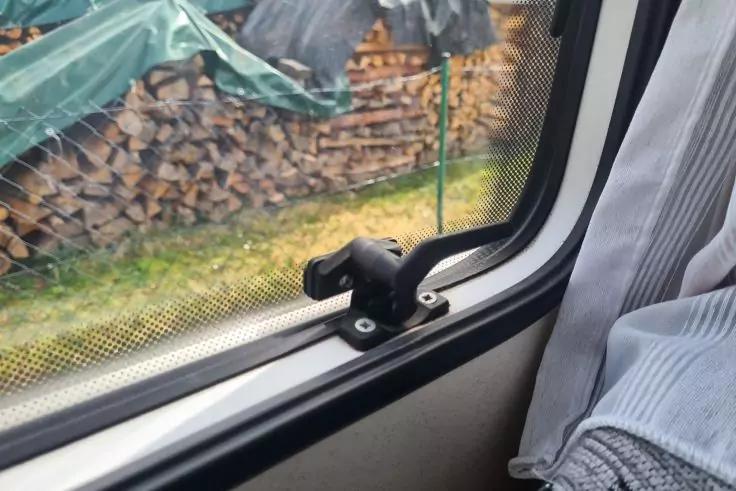 Fensterriegel am Innenrahmen, punktierte Scheibenrandbeschichtung, rechts gefalteter Vorhang, au&szlig;en gestapelte Holzscheite unter gr&uuml;ner Plane und Gras