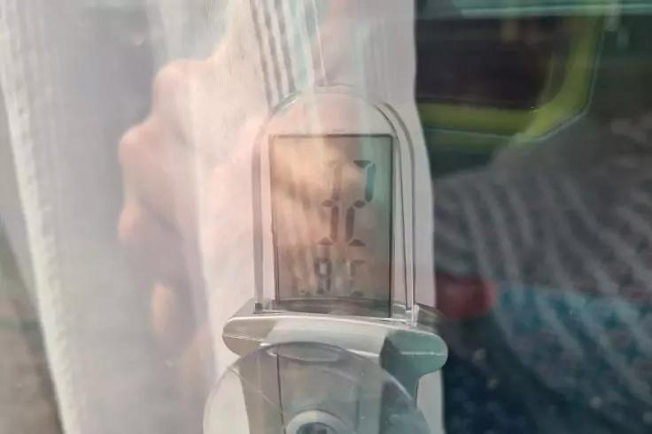 Nahaufnahme eines silbernen Metallgriffs mit einem rechteckigen transparenten Sichtfenster, in dem eine Skala mit Zahlen sichtbar ist; Reflexion einer Hand, gestreiftes Kleidungsst&uuml;ck und Vorhang im Hintergrund