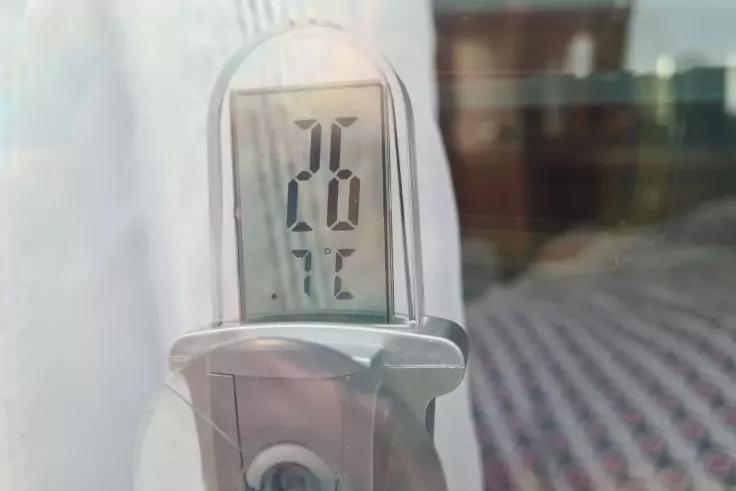 Digitales Thermometer vor einer Glasscheibe, Display zeigt gro&szlig;e Ziffern '26' und '&deg;C', links wei&szlig;er Vorhang, im Hintergrund Teppichmuster