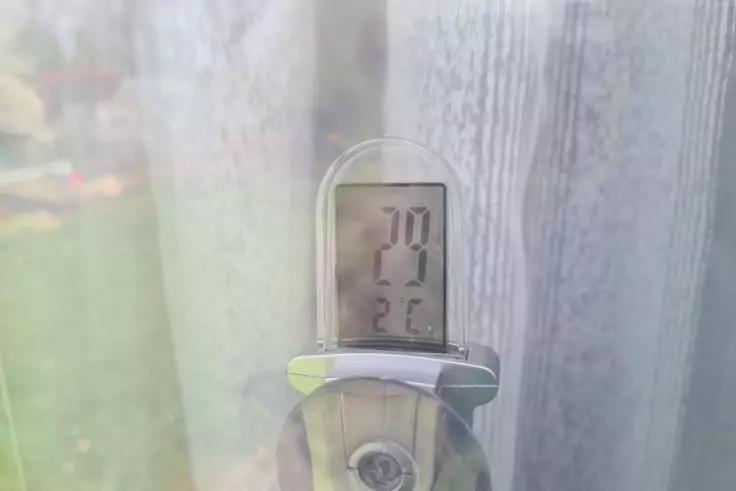 Kleines Ger&auml;t mit digitalem Display, Anzeige '27 &deg;C', befestigt mit Saugnapf an einer Fensterscheibe; dahinter Vorhang und unscharfer Garten