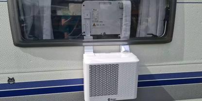 Wei&szlig;es rechteckiges L&uuml;ftungsger&auml;t in ge&ouml;ffnetem Fenster eines Wohnwagens; Gardinen im Fenster, hellgraue Au&szlig;enwand mit zwei blauen Streifen, kleiner schwarzer Katzenaufkleber unten links