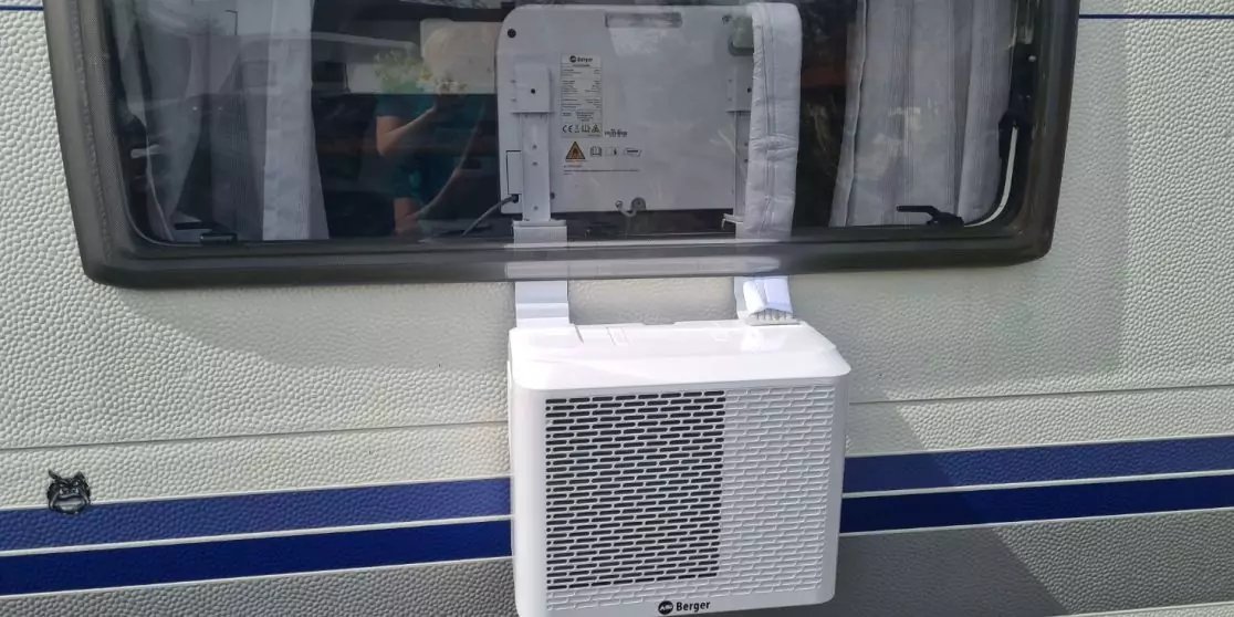 Wei&szlig;es rechteckiges L&uuml;ftungsger&auml;t in ge&ouml;ffnetem Fenster eines Wohnwagens; Gardinen im Fenster, hellgraue Au&szlig;enwand mit zwei blauen Streifen, kleiner schwarzer Katzenaufkleber unten links