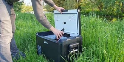 Teilweise sichtbare Person (nur H&auml;nde und Beine) &ouml;ffnet ein schwarzes, rechteckiges Ger&auml;t mit wei&szlig;em Deckel auf hohem Gras neben einem Gew&auml;sser und B&auml;umen