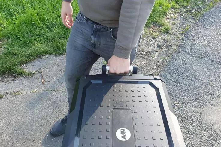 Oberk&ouml;rper und Beine einer Person, die einen schwarzen Koffer am Griff h&auml;lt, stehend auf Asphalt neben einem Grasstreifen