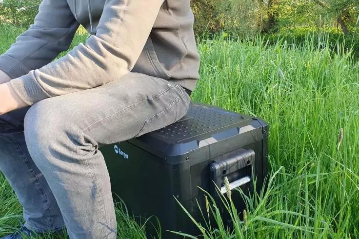 Unterer Torso und Beine einer sitzenden Person in grauem Kapuzenpullover und Jeans auf einer schwarzen Kunststoffbox mit Griff im hohen gr&uuml;nen Gras; B&auml;ume im Hintergrund