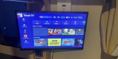 Wandmontierter Fernseher zeigt Smart‑TV‑Startbildschirm mit App‑Kacheln, darunter eine Kachel mit Aufschrift 'BBC SPORT'; rechts Gardine