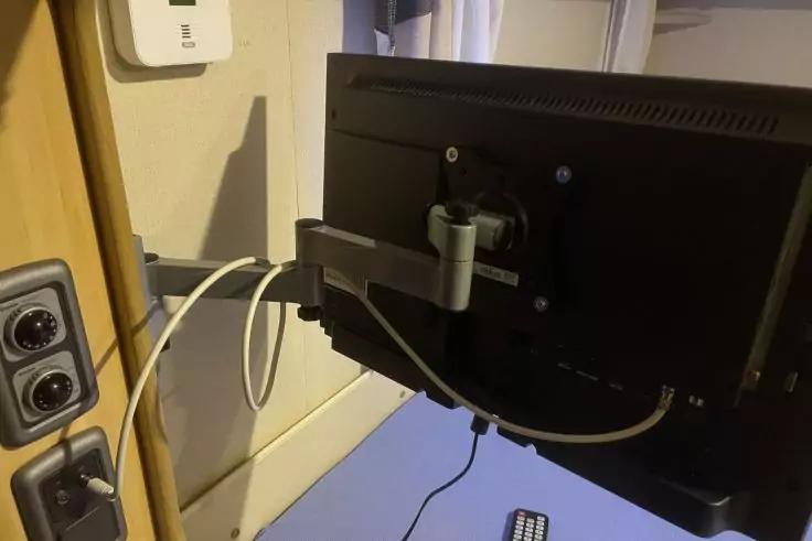 Schwarzer Monitor an einem beweglichen Monitorarm, wei&szlig;e Kabel, blauer Tisch, Fernbedienung, Steckdosen an der Wand