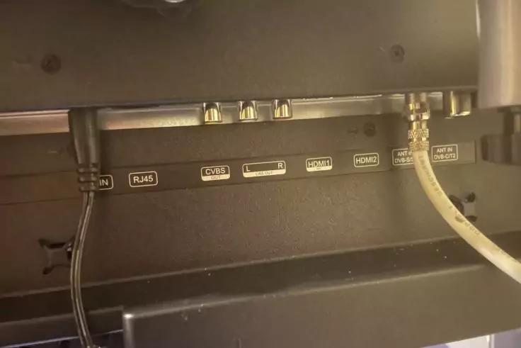 R&uuml;ckseite eines schwarzen Ger&auml;ts mit einer Reihe beschrifteter Buchsen und zwei eingesteckten Kabeln: links ein d&uuml;nnes schwarzes Kabel, rechts ein helles Kabel mit Schraubverbindung