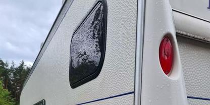 Ecke eines wei&szlig;en Wohnwagens mit get&ouml;ntem Fenster, rotem R&uuml;cklicht, blauem Streifen und Regentropfen; im Hintergrund B&auml;ume und bew&ouml;lkter Himmel
