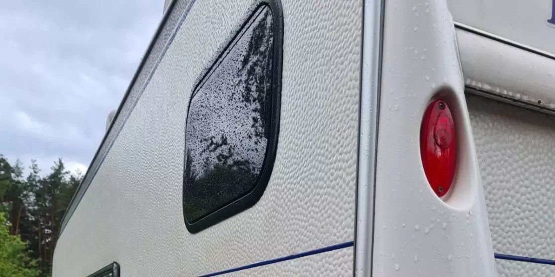 Ecke eines wei&szlig;en Wohnwagens mit get&ouml;ntem Fenster, rotem R&uuml;cklicht, blauem Streifen und Regentropfen; im Hintergrund B&auml;ume und bew&ouml;lkter Himmel