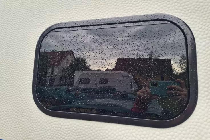 Abgerundetes rechteckiges Fenster mit Regentropfen, Spiegelung von H&auml;usern, einem Wohnwagen und einer Person, die ein Smartphone h&auml;lt