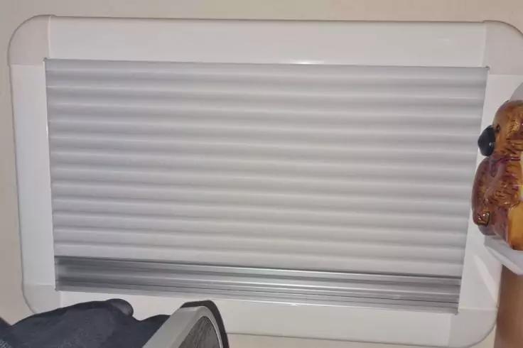 Geschlossenes wei&szlig;es Rollo im Fensterrahmen; unten links Teil einer dunklen Tasche; am rechten Rand eine braune Holzfigur