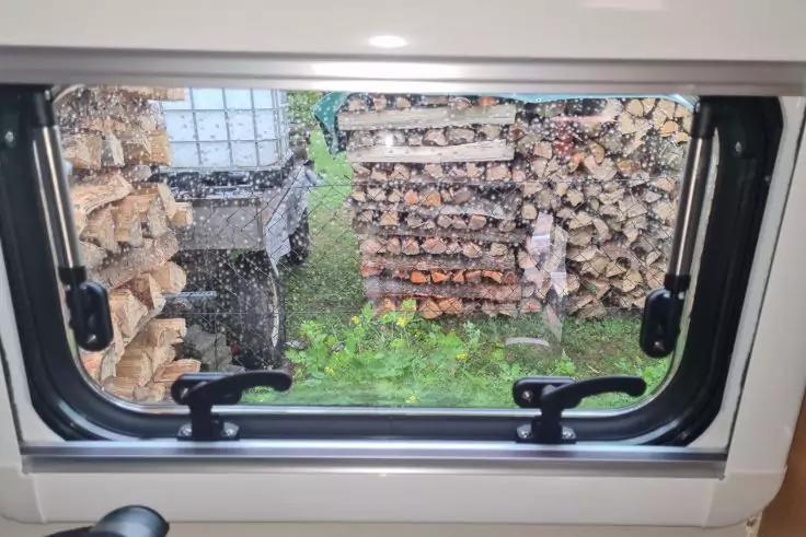 Blick durch ein Fenster mit zwei schwarzen Griffen und Regentropfen auf der Scheibe; drau&szlig;en mehrere gestapelte Holzscheite in Regalen und gr&uuml;nes Gras am Boden