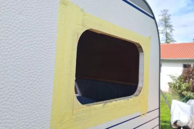 Seitenwand eines Wohnwagens mit rechteckiger Fenster&ouml;ffnung, gelbes Abklebeband um die &Ouml;ffnung, dunkles Inneres sichtbar, Hausdach und B&auml;ume im Hintergrund