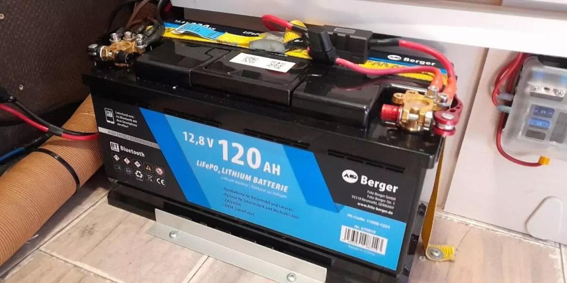 Schwarze Batterie mit blauem Etikett '12,8V 120 Ah', rote und schwarze Kabelanschl&uuml;sse, auf Metallhalterung, neben Kabeln und einer transparenten Kunststoffbox