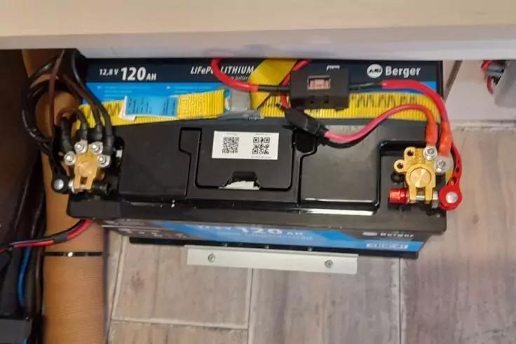 Schwarze, rechteckige Batterie mit QR-Code und Aufklebern, rote und schwarze Kabel an Polklemmen, Metallschiene auf grauem Fliesenboden, Regal mit Kabeln dar&uuml;ber