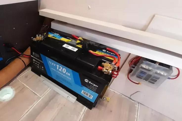 Blau-schwarze Batterie mit Aufdruck "12V 120Ah" und angeschlossenen roten sowie schwarzen Kabeln, auf Holzfu&szlig;boden vor wei&szlig;er Wand; rechts transparente Sicherungsbox mit roten Kabeln
