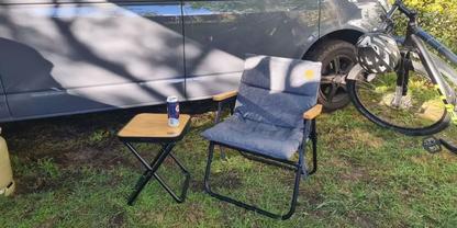 Blauer klappbarer Stuhl mit gepolstertem Sitz und kleinem rechteckigen Kissen mit gelbem Kreis; daneben ein kleiner klappbarer Beistelltisch mit einer blauen Dose; im Hintergrund die Seitenwand eines Fahrzeugs, rechts ein Fahrrad mit sichtbarem Vorde