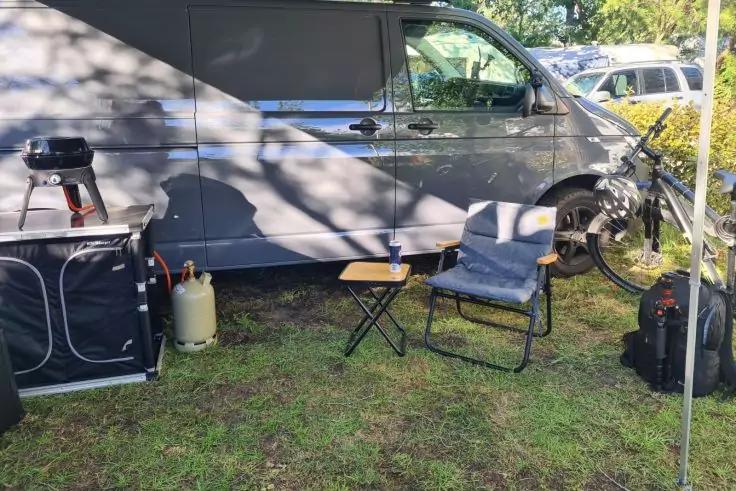 Grauer Camper-Van auf Gras mit Sonnensegel; davor ein kleiner runder Grill auf einem Klapptisch, eine Gasflasche, ein niedriger Holztisch, ein gepolsterter Klappstuhl, ein Hocker, ein Rucksack und ein Fahrrad