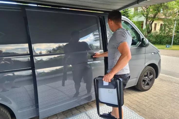 Mann steht neben grauem Transporter, h&auml;lt zusammengeklappten Klappstuhl, Schiebet&uuml;r halb ge&ouml;ffnet