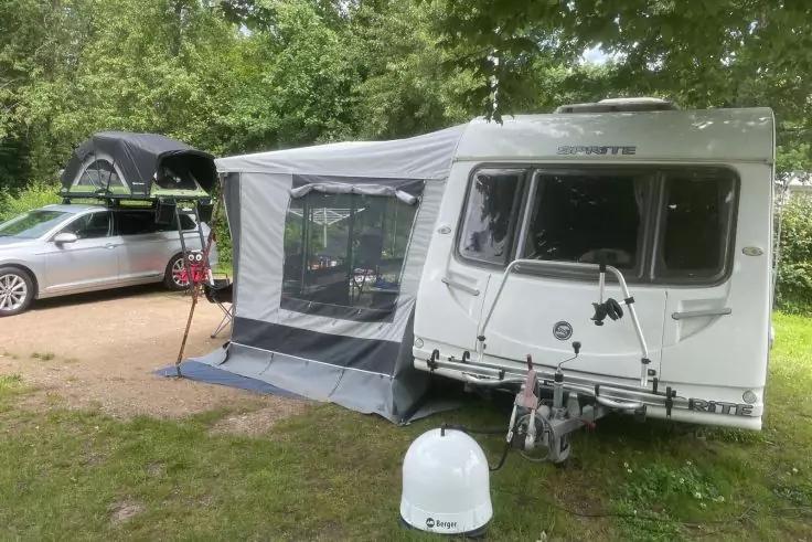 Wei&szlig;er Wohnwagen mit grauem Vorzelt und Netzfenster, daneben ein silberner Kombi mit offenem Dachkoffer, grasige Fl&auml;che und B&auml;ume, vorn eine wei&szlig;e runde Kuppel auf dem Boden
