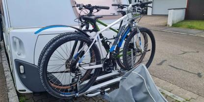 Zwei Mountainbikes auf einem Hecktr&auml;ger an einem wei&szlig;en Wohnwagen; eine graue Fahrradabdeckung liegt auf dem gepflasterten Boden daneben
