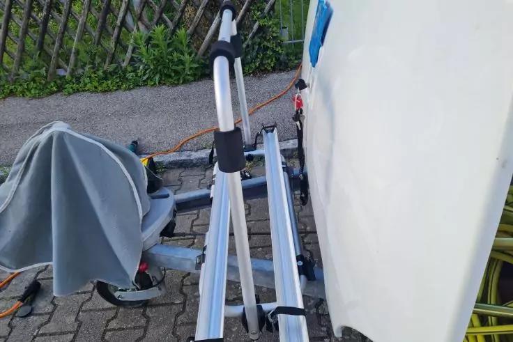 Heck eines wei&szlig;en Fahrzeugs mit silbernen Metallschienen einer montierten Halterung, schwarzem Haltearm, orangenem Spanngurt und grauer Abdeckung; gepflasterter Boden und Zaun mit Gr&uuml;n im Hintergrund