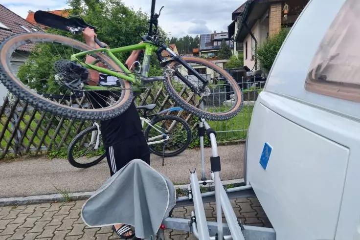 Person h&auml;lt zwei Fahrr&auml;der (gr&uuml;n, schwarz) &uuml;ber der Schulter neben einem Wohnwagen auf einem gepflasterten Weg