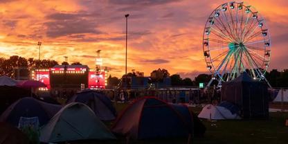 Zelte auf einer Wiese bei Sonnenuntergang, ein beleuchtetes Riesenrad und eine beleuchtete B&uuml;hne im Hintergrund