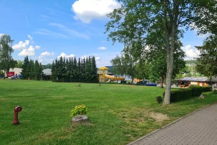 Gro&szlig;e Rasenfl&auml;che mit Spielger&auml;t (gelbe Rutsche), B&auml;ume, gepflasterter Fu&szlig;weg rechts, roter Hydrant vorne links, Geb&auml;ude im Hintergrund, blauer Himmel mit Wolken