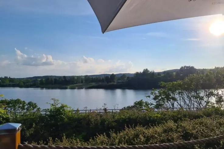 Blick auf einen See mit B&auml;umen am gegen&uuml;berliegenden Ufer, ein Sonnenschirm oben rechts, eine Font&auml;ne in der Ferne, Seil mit Pfosten und ein M&uuml;lleimer im Vordergrund