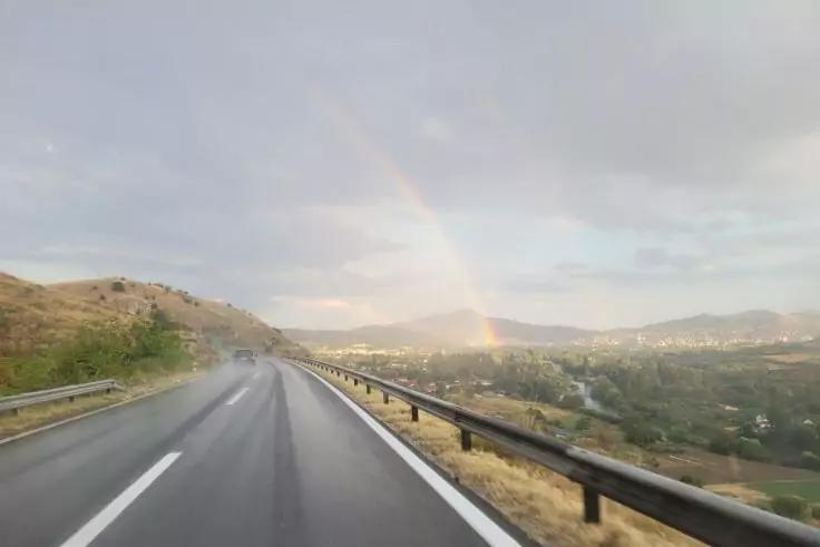 Nasse Straße mit Leitplanke, Hügeln links und Regenbogen im bewölkten Himmel über einem Tal