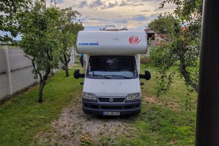 Frontansicht eines weißen Wohnmobils auf einer Grasfläche zwischen Bäumen, links ein Zaun, rechts ein Haus, Kiesweg vor dem Fahrzeug, bewölkter Himmel