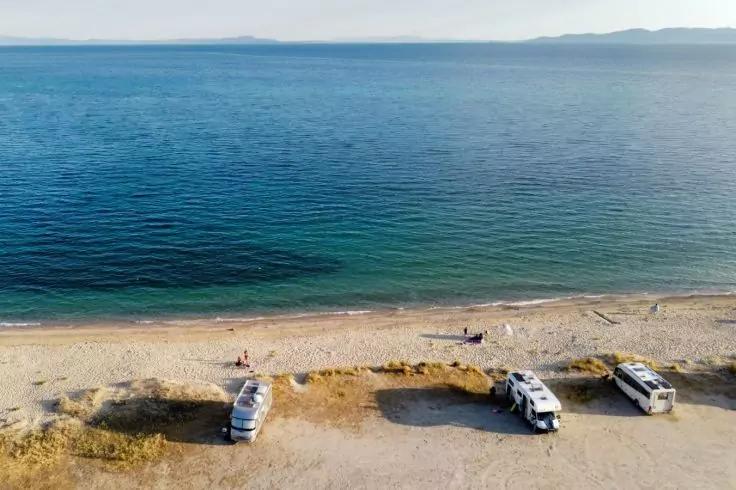 Luftaufnahme eines sandigen Strandes mit drei Wohnmobilen am Ufer, wenigen Personen und zwei Kajaks am Rand des blauen Meeres; Land am Horizont