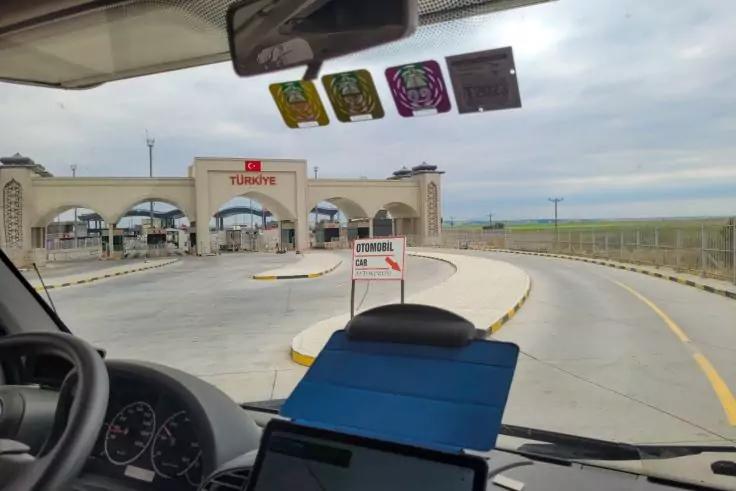 Blick aus dem Fahrzeug über Lenkrad und Armaturenbrett mit Tablet auf eine gebogene Straße zu einem Gebäude mit drei Bögen; Windschutzscheibe oben drei Aufkleber