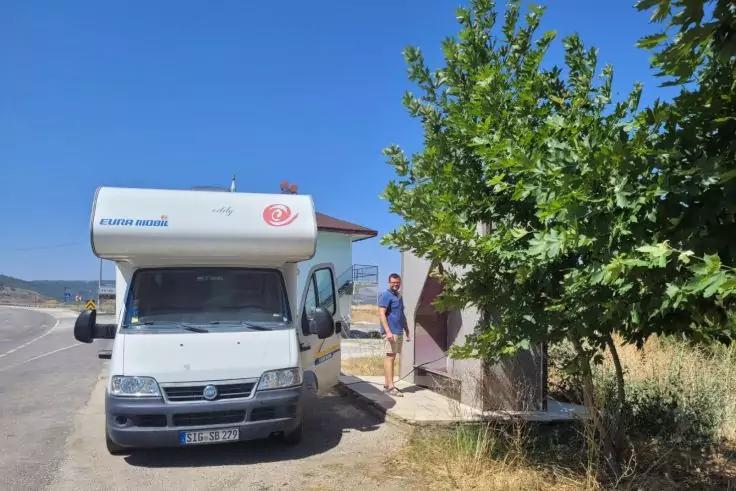 Weißes Wohnmobil am Straßenrand; eine Person steht neben dem Wohnmobil vor einem kleinen Gebäude; rechts ein grüner Baum; blauer Himmel