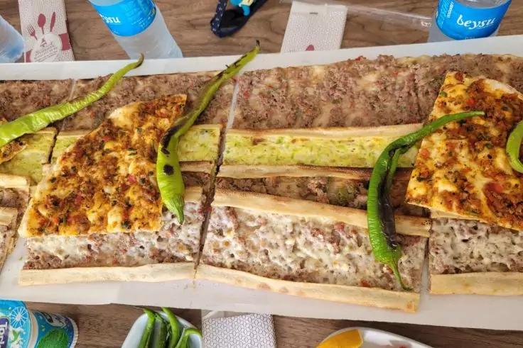 Drei längliche Fladenbrotstücke mit Belag auf einem Holzbrett, jeweils grüne Chilischoten darauf, Tisch mit Wasserflaschen und Servietten im Hintergrund