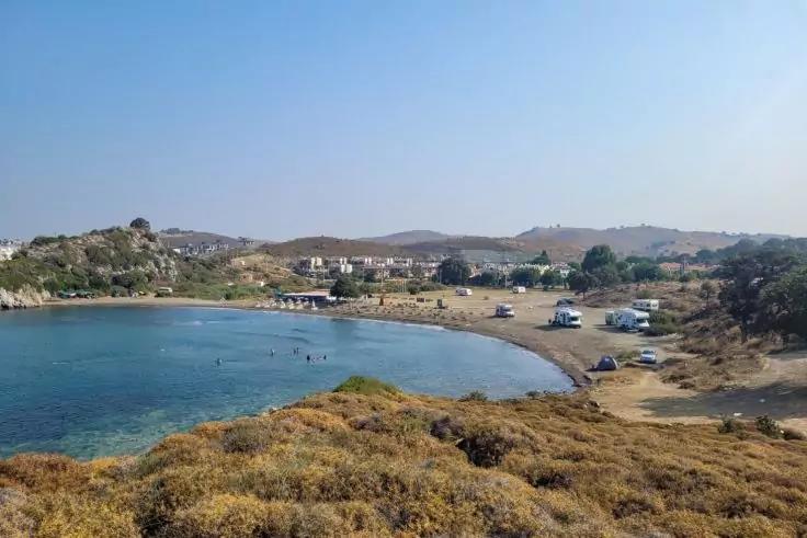 Kleine Bucht mit türkisfarbenem Wasser und Strand, Personen im Wasser, Wohnmobile und Zelte am Ufer, trockene Büsche im Vordergrund, niedrige Hügel und Häuser im Hintergrund, blauer Himmel