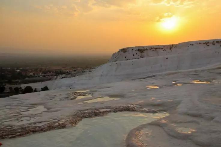 Weiße, terrassenförmige Becken mit flacher Wasseroberfläche, im Hintergrund ein orangefarbener Sonnenuntergang über einer flachen Landschaft