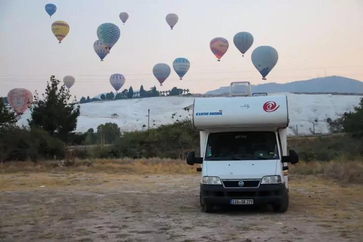 Wohnmobil auf unbefestigtem Boden, dahinter Bäume und eine helle weiße Fläche, mehrere Heißluftballons am Himmel, entfernte Hügel