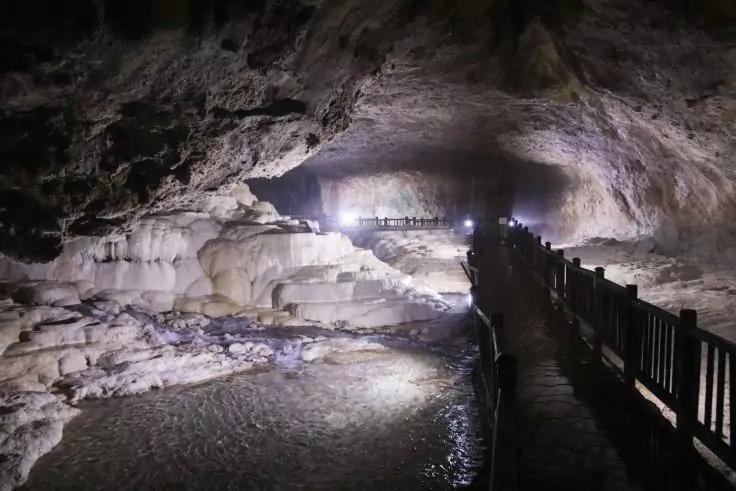 Innenraum einer Höhle mit beleuchtetem Gewölbe, weißen terrassenartigen Mineralablagerungen und flachem Wasser links, hölzerner Steg mit Geländer rechts, mehrere Personen als dunkle Silhouetten auf dem Steg, Lichter und Lichtreflexe im Wasser