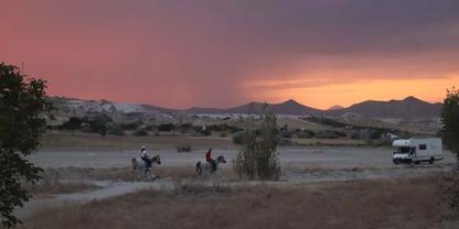 Personen auf Pferden auf einem Feldweg, Wohnmobil rechts, trockene Wiese, H&uuml;gel und rosa-oranger Himmel